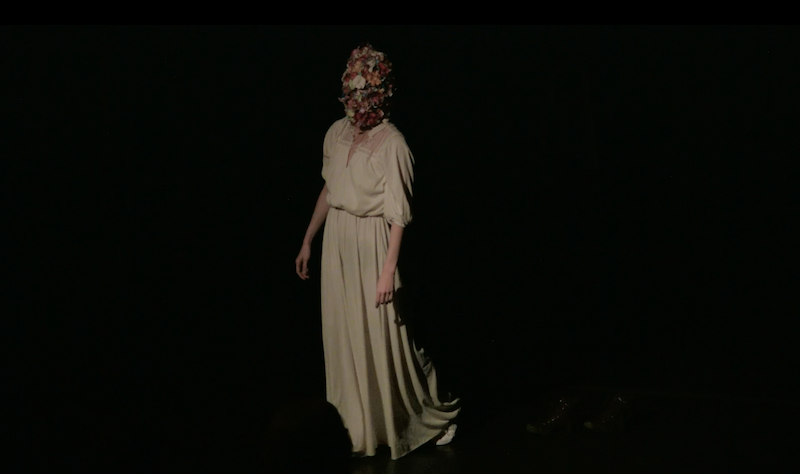 A woman in a white Edwardian dress wears a mask covered in flowers.